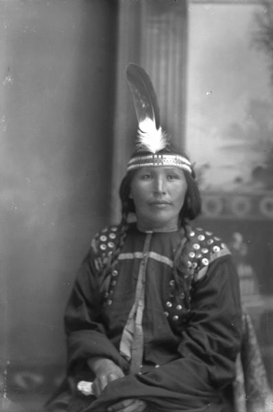 Studio portrait of Nora Arthur, Greengrass, sitting and wearing regalia, including a traditional blouse, hair braids, and a beaded headband with an eagle feather. In the background is a painted backdrop.