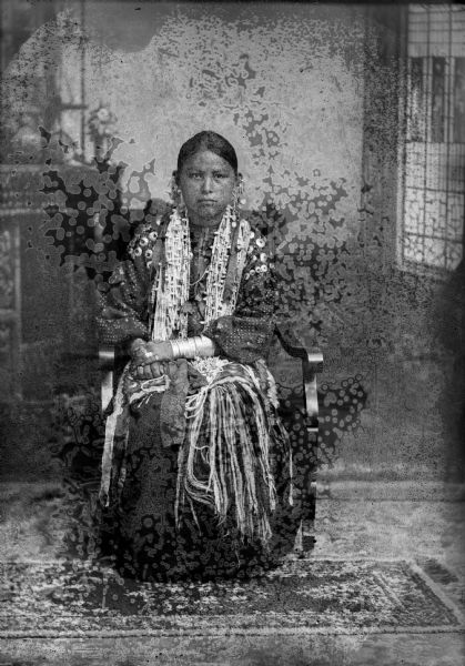 Studio portrait in front of a painted backdrop of a Ho-Chunk girl posing sitting and wearing several necklaces, file bracelets, earrings, rings, and a traditional blouse and skirt.