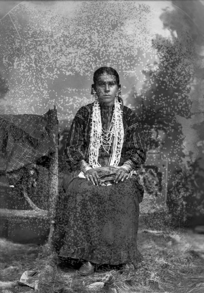 Studio portrait in front of a painted backdrop of a Ho-Chunk girl posing sitting near a prop wood fence, draped with a plaid fringed shawl. She is wearing several necklaces, file bracelets, earrings, rings, and a traditional blouse and skirt.