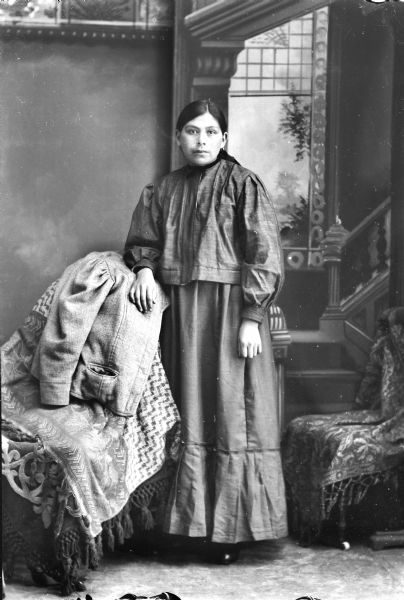 Studio full-length portrait of a Ho-Chunk woman posing in front of a painted backdrop. She is standing with her right arm on a chair which is draped with a winter coat, and she is wearing a contemporary blouse and skirt.