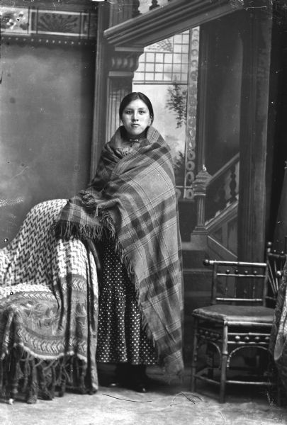 Studio full-length portrait of a Ho-Chunk woman in front of a painted backdrop posing standing. She is wearing a dark polka dot dress and is wrapped in a plaid fringed shawl. There are chairs on either side of her, and the chair to the left is draped with fabric.