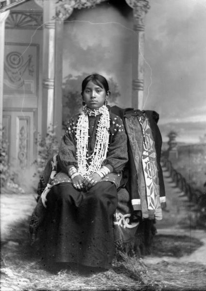 Studio full-length portrait in front of a painted backdrop of a Ho-Chunk girl posing sitting and wearing regalia, including several necklaces, earrings, file bracelets, and rings. She is sitting on a chair draped with a ribbon work shawl.