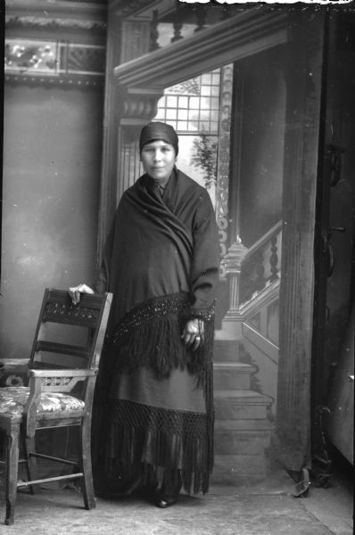 Studio full-length portrait in front of a painted backdrop of a Ho-Chunk woman posing standing with her right hand on a chair. She is wearing a head scarf, and is wrapped in a fringed shawl.