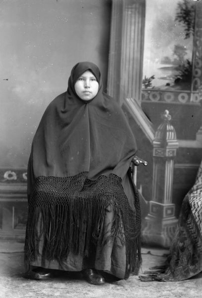 Studio full-length portrait of a seated Ho-Chunk woman. She is wrapped in a fringed shawl, showing only her face. Her name is Annie Lizzie Falcon, Whitedog, Brown, Hall (Das Chunt A Win Kah) (wife of William Hall) (daughter of Thomas Falcon [Cho Ne Mon E Kah] and Lucy Whitespot [Kee Ree Jay Win Kah]). In the background is a painted backdrop.

