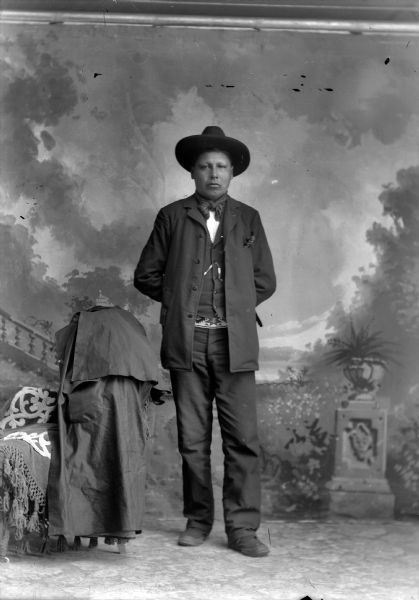 Studio full-length portrait in front of a painted backdrop of a Ho-Chunk man posed standing with his hands behind his back. He is wearing a suit, bandana, hat, and beaded belt. To the left of him is a chair draped in fabric and a coat (duster).