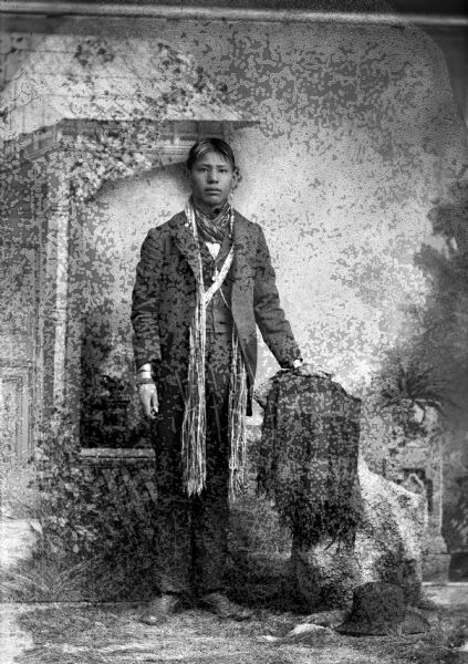Full-length studio portrait of Frank C. Thunder (son of [Wa Con Cha Kah] John Thunder aka Dr. Thunder and [We Hon Pe Kaw] Lucy Bear, Thunder). He is standing next to a stump draped with a plaid shawl/blanket and a hat on the ground. He is wearing a suit, long earrings, long streamers, a sash, and file bracelets. In the background is a painted backdrop.