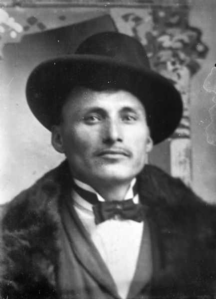 Copy photograph of a studio head and shoulders portrait of a Ho-Chunk man wearing a winged-collar shirt, bow tie, fur collar coat, and hat.