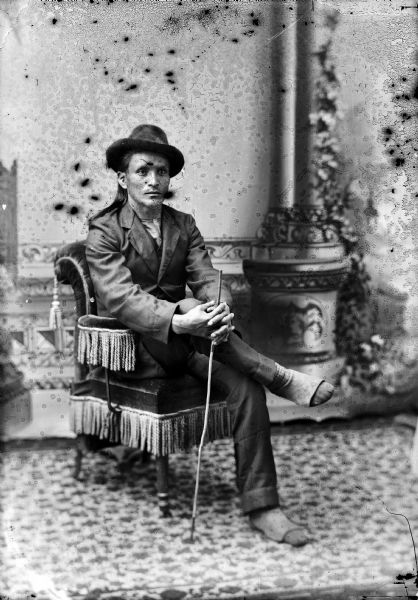 Studio full-length portrait in front of a painted backdrop of a Ho-Chunk man with long hair posed sitting in a fringed chair, holding a stick in clasped hands over his crossed legs. He is wearing a suit, hat, and moccasins.