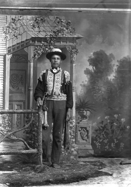 Studio full-length portrait in front of a painted backdrop of Frank Winnesheik posing standing. He has his right hand on a prop wooden fence, and is wearing a beaded shirt, gorget, hat, and woven sash.