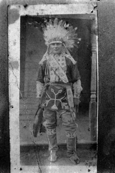 Copy photograph of a Ho-Chunk man posed standing outside a building wearing a Sioux Bonnet, woodland design breech cloth, beaded shirt, bandoleer, and garters.