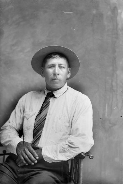 Three-quarter length studio portrait of Edward Winneshiek sitting with his hands folded, wearing a light-colored shirt, necktie, and hat.


