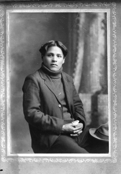 Copy photograph of a Ho-Chunk man posed sitting with his hands clasped, a hat on his lap, wearing a suit coat and turtle neck sweater.