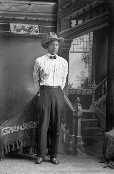 Studio full-length portrait in front of a painted backdrop of a Ho-Chunk man posed standing with his hands behind his back, wearing a light-colored shirt, a bow tie, and hat.