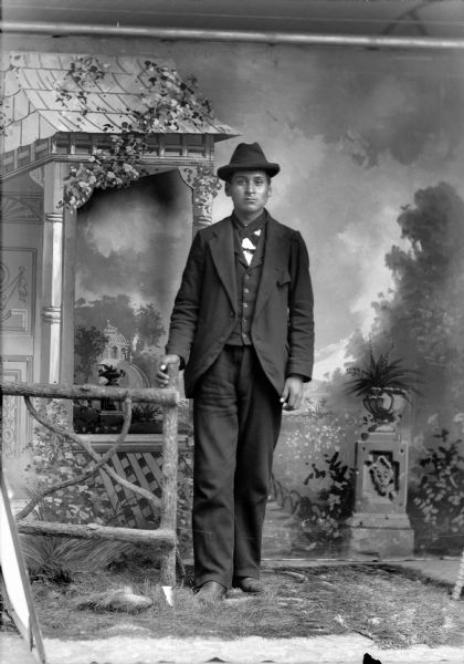 Studio full-length portrait in front of a painted backdrop of a Ho-Chunk man posed standing with his right hand on a prop wooden fence and wearing a suit, bandana, and hat.