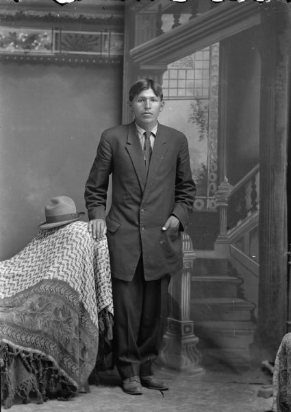 Studio full-length portrait in front of a painted backdrop of a Ho-Chunk man posed standing with his right hand on a draped chair that also holds a hat. His left hand is in his long suit coat's pocket. He is also wearing a necktie.