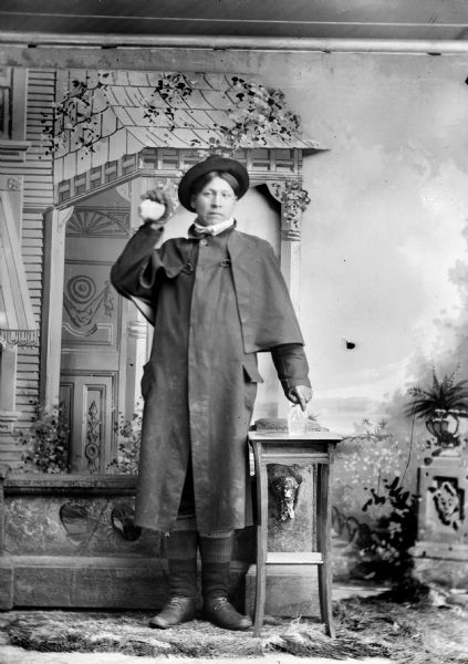 Studio full-length portrait in front of a painted backdrop of a Ho-Chunk man posed standing wearing an overcoat (duster) and hat. He is holding a spherical object (snowball?) in his raised right hand, and with his left hand is holding onto a liquor bottle which is on a table in front of a prop stone wall.