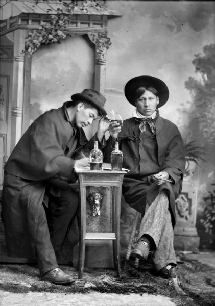 Studio portrait in front of a painted backdrop of a Ho-Chunk man posed sitting on a prop fence on the left writing a letter on a small table with his right hand, and his left hand on the brim of his hat. The man on the right is posed sitting, holding a liquor bottle in his right hand and a cork in his left hand. Both men wear long coats (dusters) and hats. On the table are two liquor bottles.