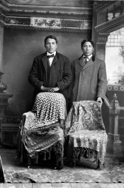 Studio portrait in front of a painted backdrop of two Ho-Chunk men posed standing behind chairs draped with fringed cloth. The men are both wearing suits and coats. The man on the left wears a bow tie and the man on the right wears a necktie.