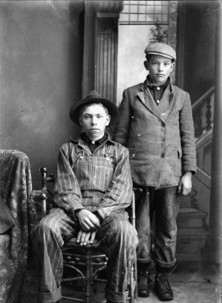 Studio portrait in front of a painted backdrop of a European American boy posed sitting on the left, wearing striped coveralls, a striped shirt, and a hat. Another European American boy is posed standing on the right wearing a suit coat and cap.