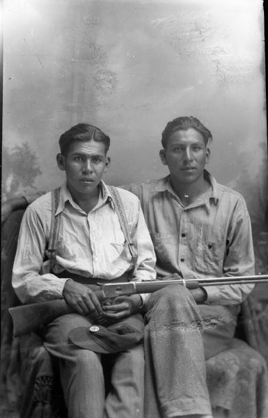 Studio portrait in front of a painted backdrop of two Ho-Chunk men posed sitting next to each other. The man on the left is wearing suspenders and holding a rifle and a cap on his lap. The man on the right has his right arm behind the other man. Both men wear light-colored shirts and dark-colored pants. A button or emblem on the cap appears to be a 4-H symbol.