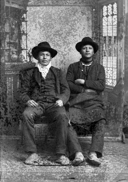 Studio portrait in front of a painted backdrop of two Ho-Chunk men posed sitting. The man on the left is wearing a suit, bandana, and a hat, and the man on the right has his arms crossed and is wearing a hat, short necklace, and an overcoat.