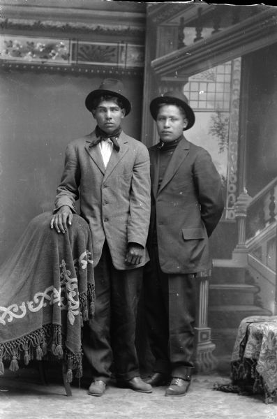 Studio full-length portrait in front of a painted backdrop of two Ho-Chunk men posed standing and wearing suits and hats. The man on the left has his right hand on a chair draped with a fringed throw, and is wearing a light-colored shirt and bandana. The man on the right has his hands behind his back and is wearing a turtleneck sweater.