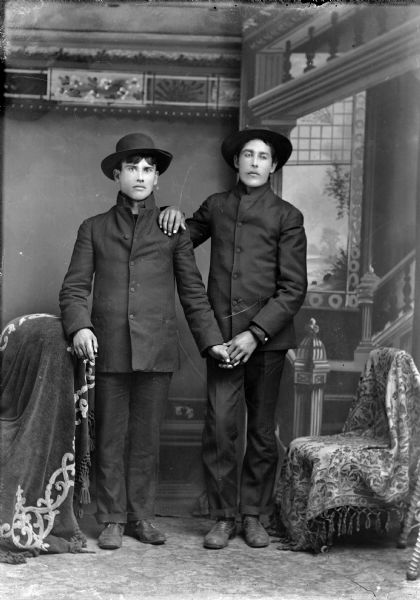 Studio full-length portrait in front of a painted backdrop of two young Ho-Chunk men posed standing near a chair draped with a fringed throw. The man on the left clasps his left hand in the left hand of the man on the right, and the man on the right has his right hand on the other man's left shoulder. Both are wearing dark suit coats and hats.