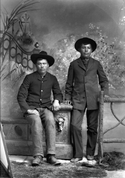 Studio full-length portrait of two Ho-Chunk men posed in front of a painted backdrop. The man on the left is sitting on a prop stone wall and another Ho-Chunk man is standing on the right resting his hand on the pillar and his other hand on a prop wooden fence post. Both men are wearing suits and hats, and the man on the right also wearing a bandana.