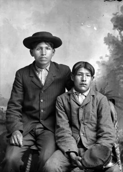 Studio portrait in front of a painted backdrop of two Ho-Chunk boys posed sitting wearing suits and striped shirts. The boy on the left is sitting on a stool or table and is wearing a hat. The boy on the right is holding a cap.