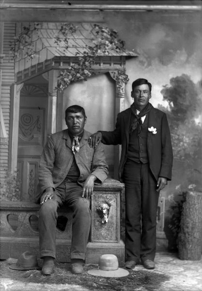 Studio portrait in front of a painted backdrop of a Ho-Chunk man posed sitting on the left on a prop stone wall, and Henry Winneshiek (ni do ni KA) on the right, standing with his right hand on the left shoulder of the sitting man and is wearing earrings and a ring. Both men are wearing suits and bandanas and have hats near their feet on the ground.