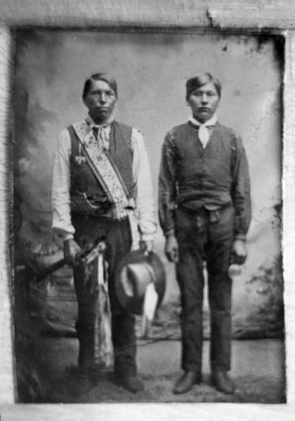 Copy photograph of a studio portrait of two Ho-Chunk men posing standing. The man on the left is wearing a dark vest, light-colored shirt, bandana, and bandoleer, and holding an object in his right hand and a hat in his left hand. The man on the right is wearing a dark-colored shirt and vest, and a light-colored bandana.