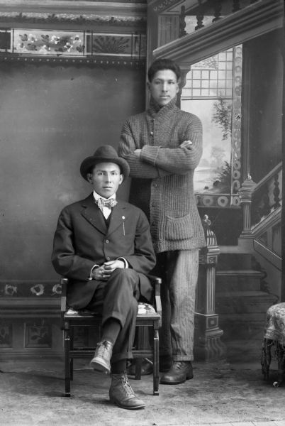 Studio portrait in front of a painted backdrop of a Ho-Chunk man posed sitting on the left with his legs crossed, hands clasped, and wearing a suit, bow tie, and hat, and a European American man posed standing on the right with his arms crossed and wearing a long button-down sweater.