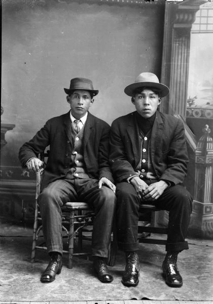 Studio portrait in front of a painted backdrop of two young Ho-Chunk men posed sitting and wearing suits and hats. The man on the left also wears a necktie, and the man on the right is identified as probably Robert Greengrass.
