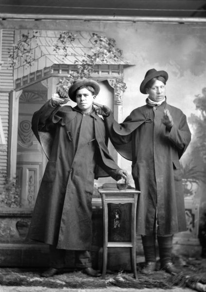 Studio full-length portrait in front of a painted backdrop of two Ho-Chunk men posed standing and wearing overcoats (dusters), gloves, and hats. The man on the left is holding a liquor bottle up in his right hand and another bottle on a small table with his left hand. The man on the right is pointing at the other man with his right hand and holding something in his left hand.