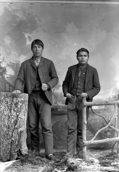 Studio full-length portrait of two Ho-Chunk men standing. The man on the left has his right hand on a stump and the man on the right, Frank Yellowfeather Climer, has his left hand on a prop wooden fence post. Both men are wearing suits and bandanas. In the background is a painted backdrop.