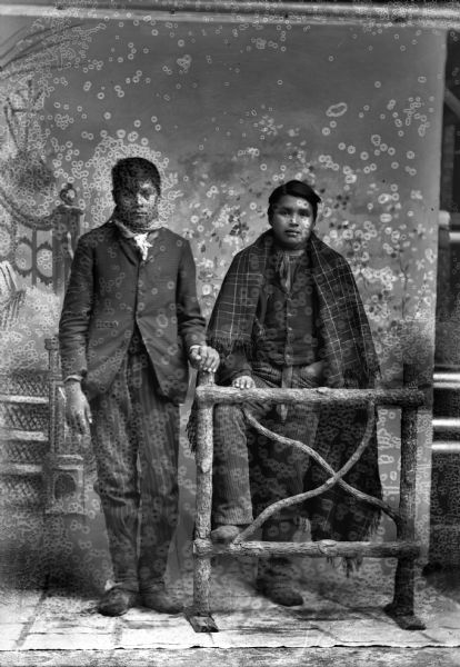 Full-length studio portrait of two men. The unknown man on the left has his left hand on a prop wooden fence post, and Frank Yellowfeather Climer, on the right behind the wooden fence, is wearing a plaid fringed shawl over his shoulders. Both men wear suits and bandanas. In the background is a painted backdrop.