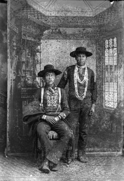 Studio portrait in front of a painted backdrop of a Ho-Chunk man posed sitting on the right with his right hand over his left hand on his lap, with a suit coat over his right arm, and wearing a ribbon work shirt and hat. Another Ho-Chunk man is posed standing on the right and is wearing a traditional shirt, vest, and hat.