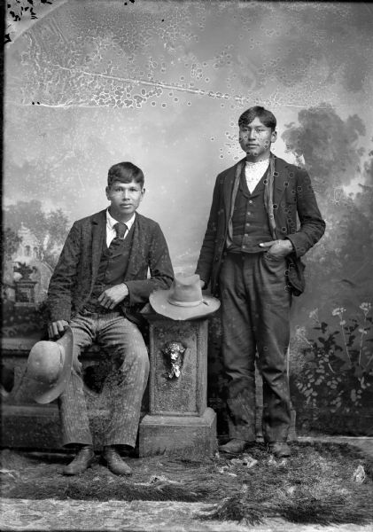 Studio portrait in front of a painted backdrop of James Seymour (Nebraskan Winnebago) sitting on a prop stone wall, holding a hat in his right hand, and wearing a suit and necktie. Frank Redcloud (Ho-Chunk) is standing, holding a hat with his right hand and resting it on the stone wall, his left hand in his pocket, and wearing a suit.