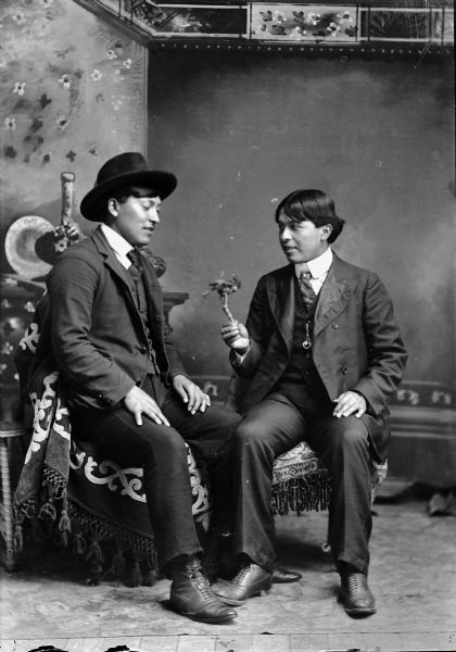 Studio portrait in front of a painted backdrop of two Ho-Chunk men posed sitting on chairs draped with a fringed throw. They are both wearing suits and neckties. The man on the left is wearing a hat and the man on the right is holding a stick with a mounted stuffed bird on it with his right hand.