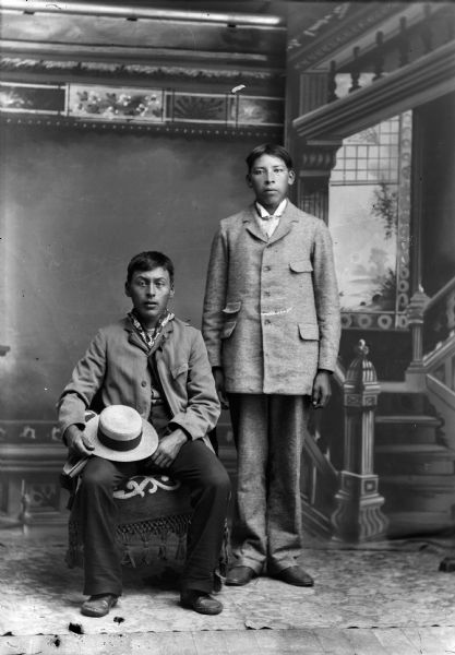 Full-length studio portrait of George Otter (Hay Cho Kah) sitting on the left in a chair draped with a fringed throw, holding a hat on his lap. George Sine is standing on the right. Both are wearing light-colored suits. In the background is a painted backdrop.