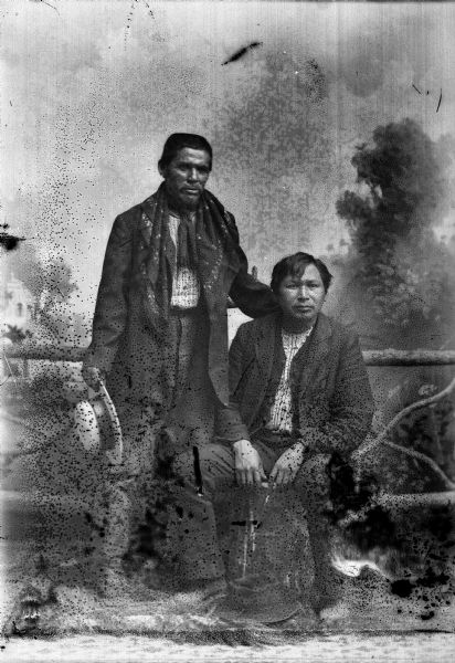 Studio portrait of Charles Stacy (Ho-Chunk) in front of a painted backdrop and prop wooden fence. He is wearing a small shawl over his shoulders and has his left hand on the right shoulder of Yellow Cloud (Nebraskan Winnebago) sitting on the right. Both men are wearing suits and have hats.