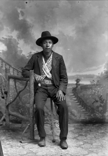 Full-length studio portrait of Henry Greencrow (Coo Noo Zee Kah) (son of [Ka He Cho Kah] Green Crow and Keeps on Moving Woman) posed sitting on a stool with his right arm resting on a prop wooden fence. He is wearing a suit, bandoleers, beaded belt, and hat. In the background is a painted backdrop.