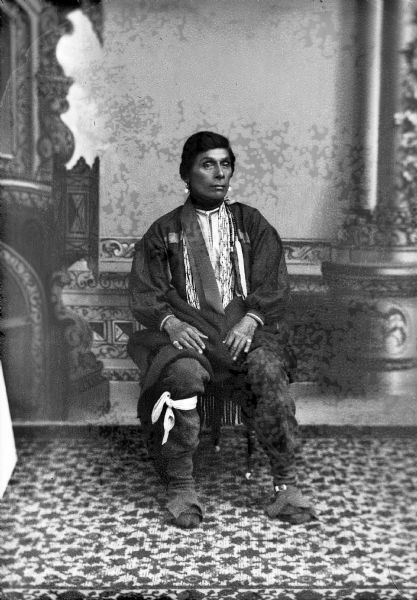 Studio portrait in front of a painted backdrop of a Ho-Chunk man posing sitting with a shawl around his waist and lap, and wearing a ribbon sash, earrings, moccasins, rings, and several necklaces. Not an original Van Schaick because of studio props.