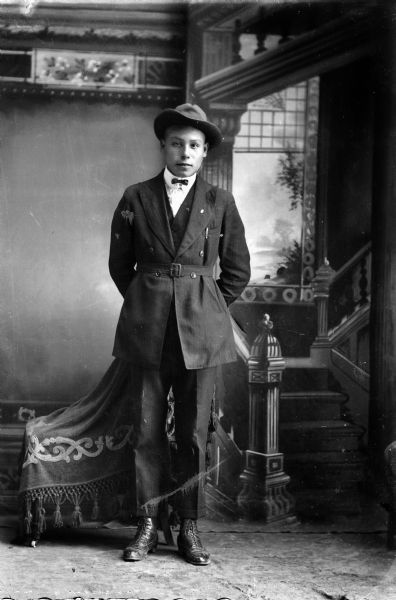 Studio full-length portrait in front of a painted backdrop of a young Ho-Chunk man posing standing with his hands behind his back. He is wearing a belted suit coat, bow tie, and hat.