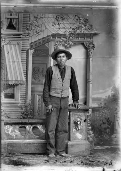 Full-length studio portrait of a Ho-Chunk man (Ae'n tto Ao K ni K K [old Ho-Chunk writing]) standing in front of a prop stone wall with his left hand on the post. He is wearing drop earrings, a dark-colored shirt, a light-colored vest, and a hat. In the background is a painted backdrop.