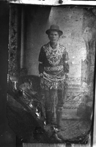 Copy photograph of a studio portrait in front of a painted backdrop of a Ho-Chunk man posing standing with his hands by his sides. He is wearing woodland-style regalia, including a beaded belt, shirt, several necklaces, and earrings.