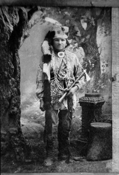Copy photograph of a full-length studio portrait of a standing Ho-Chunk man. He is holding a metal tomahawk pipe, and is wearing a bandoleer, gorget, several necklaces, and a hat with a fur tail. He is standing near a prop tree, tree stump, and pillar, with a painted backdrop behind.