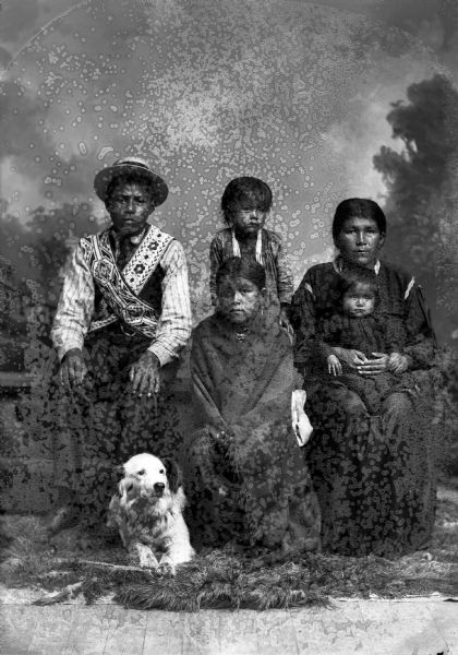 Studio portrait in front of a painted backdrop of a young Ho-Chunk man posing sitting on the left on a prop stone wall and wearing bandoleers, a necktie, and a straw hat. In the center are two seated Ho-Chunk girls: the older girl in front wrapped in a fringed shawl, and the girl behind her sitting on the stone wall is wearing several necklaces. Posing on the right is a woman sitting on the stone wall holding a small child on her lap. A light-colored dog is lying on the ground between the young man and girls.