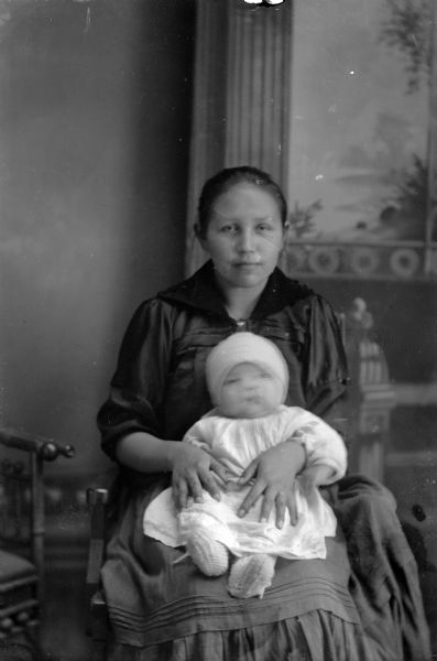 Studio portrait in front of a painted backdrop of a Ho-Chunk woman posing sitting and wearing a dark-colored dress, and holding an infant in her lap who is wearing a light-colored dress and bonnet. The infant's face is blurred from motion.
