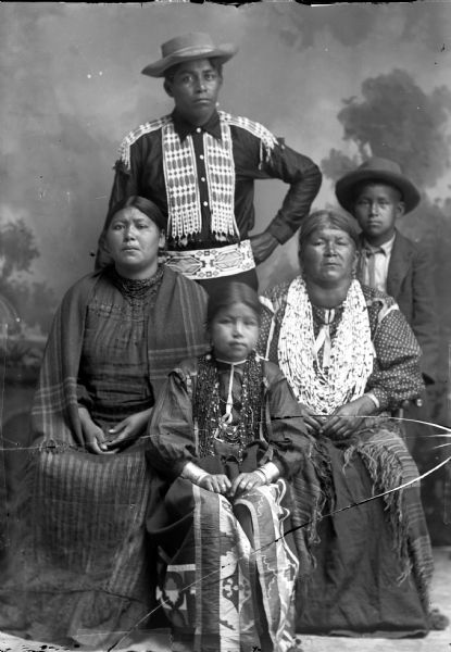 Studio portrait in front of a painted backdrop of two Ho-Chunk women posing sitting and wearing several necklaces, and a Ho-Chunk girl sitting in between them wearing several necklaces and a ribbon-work skirt. A Ho-Chunk man is posing standing behind them wearing a beaded breast and shoulder plate and a hat, and a Ho-Chunk boy is posing standing to his right wearing a bow tie and hat.
